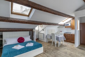 Ein Schlafzimmer mit Bett, Fenster, Dachfenster, einer Küchenzeile inklusive Mikrowelle und Holzbalken.