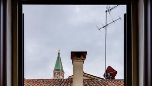 En vy genom ett fönster som visar en byggnad med ett tak täckt av tegelpannor, en skorsten och ett torn med ett grönt tak.