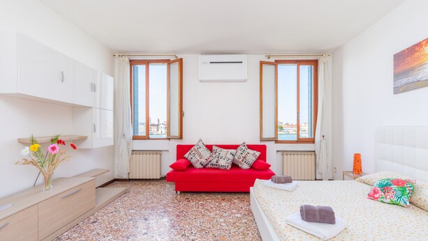 Quarto com um sofá vermelho, uma cama com roupa de cama floral, vista da cidade por grandes janelas e um ar condicionado branco.