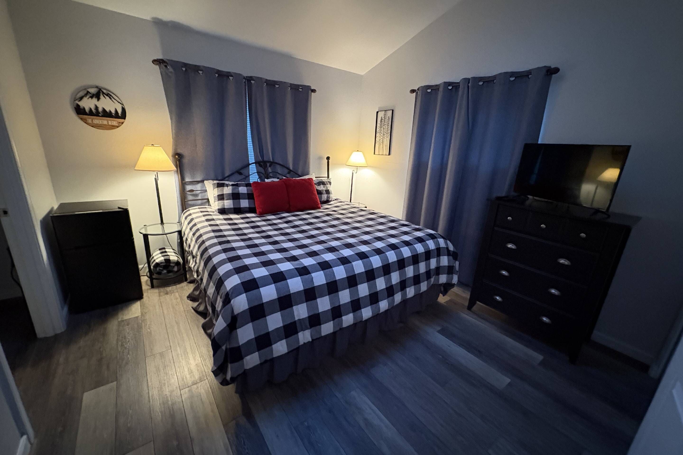 A bedroom with a checkered bedspread, a red pillow, two bedside lamps, a TV on a dresser, and a small table with a lamp.