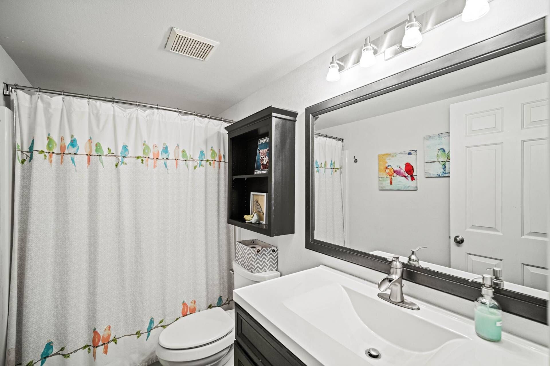 Ein Badezimmer mit einer Duschvorhang, der bunte Vogelmotive zeigt, einer weißen Toilette, einem weißen Waschbecken mit Spiegel darüber und einem Regal mit einem Schild mit der Aufschrift 'Waschen' und einem Schild mit der Aufschrift 'Butterblume'.