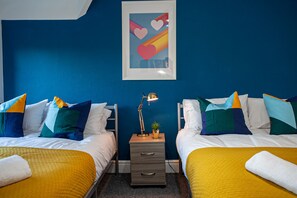 A hotel room with two beds, each with a yellow bedspread, white pillows, and colorful pillows. There is a small nightstand with a lamp and a plant. A framed picture hangs on the wall above the beds. - The Manchester Pad - Sleeps 12 (Manchester)