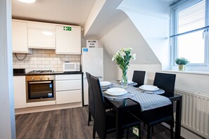 A modern kitchen with white cabinets, a built-in oven, and a microwave. There is a dining table with chairs and a vase of flowers. A sign on the wall reads "WELCOME STAY HAPPY". - The Manchester Pad - Sleeps 12 (Manchester)