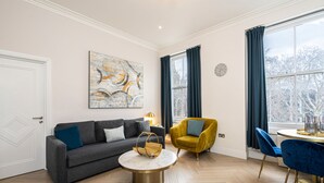 Een moderne woonkamer met een grijze bank, een ronde salontafel, een gele fauteuil en een eettafel met blauwe stoelen.