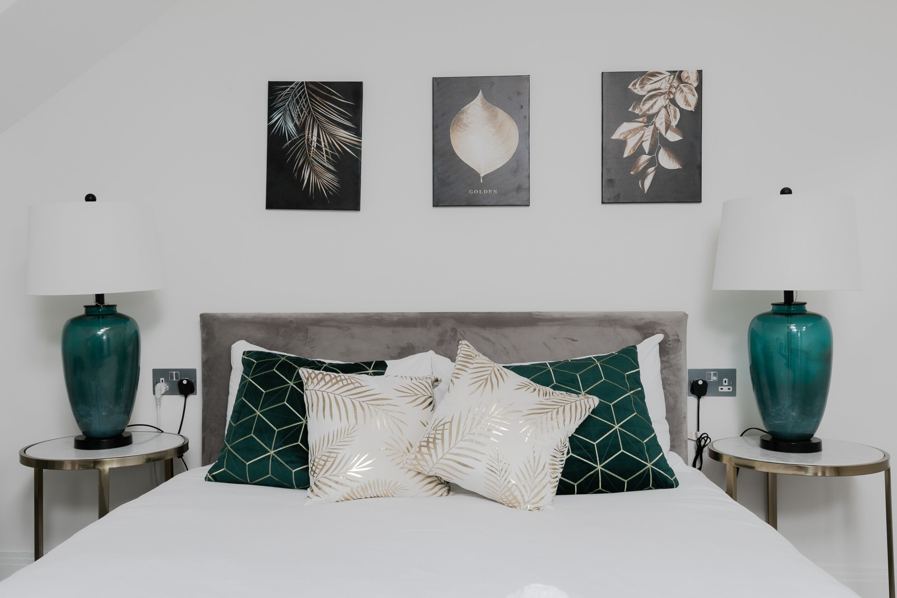 A bedroom with a bed, two bedside tables, two wall-mounted lamps, and three framed botanical prints.
