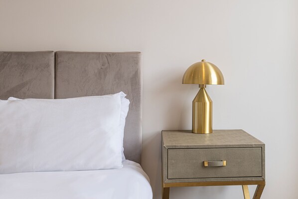 A bed with a grey headboard, a white pillow, and a bedside table with a gold lamp and a drawer.
