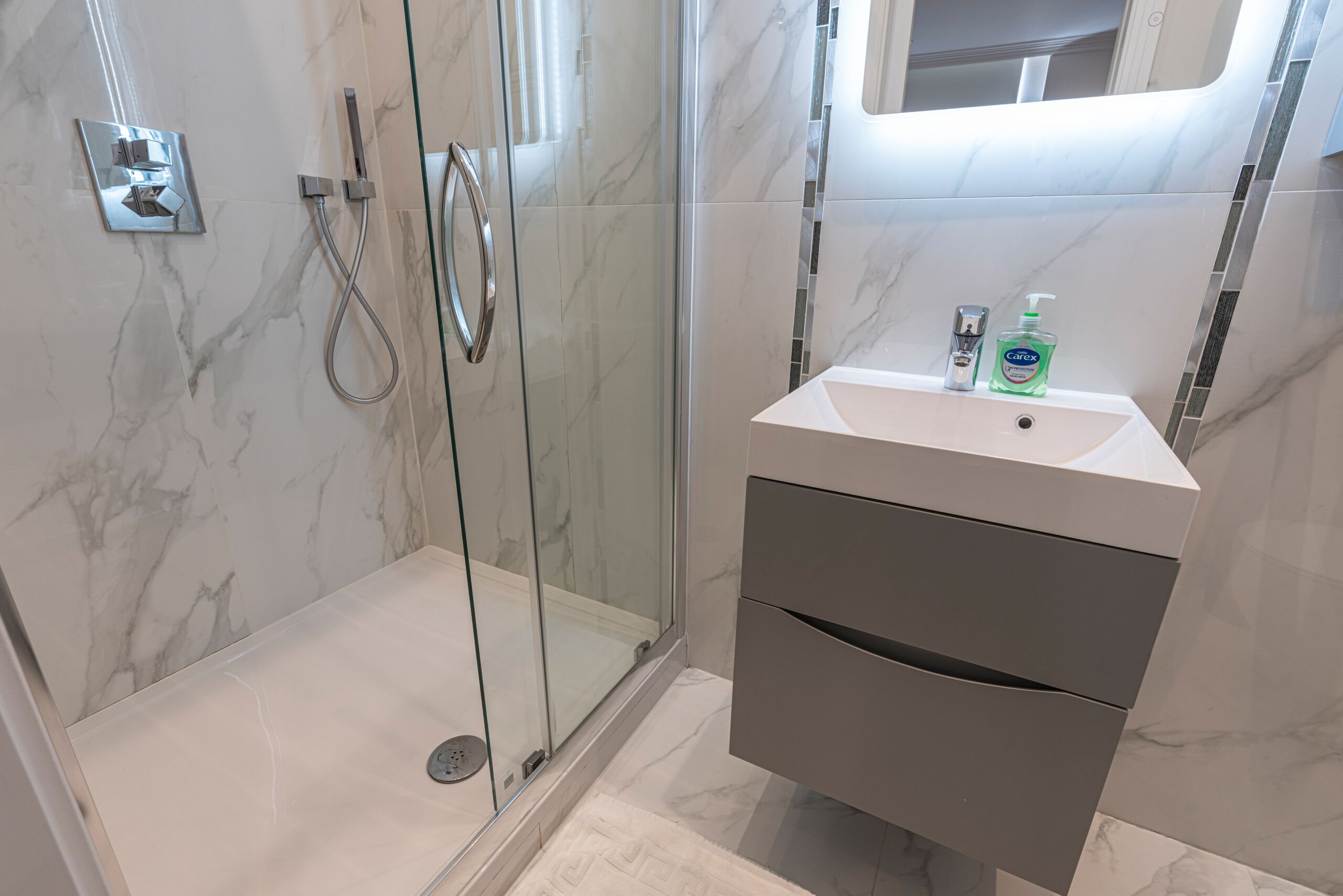 A modern bathroom with a glass shower enclosure, a marble-tiled wall, and a white sink with a soap dispenser.