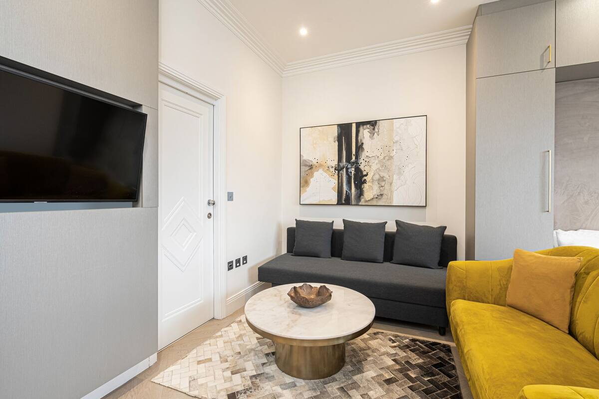 Ein modernes Wohnzimmer mit einer Couch, einem gelben Sofa, einem Couchtisch mit einer Schale und einem an der Wand montierten Flachbildfernseher.