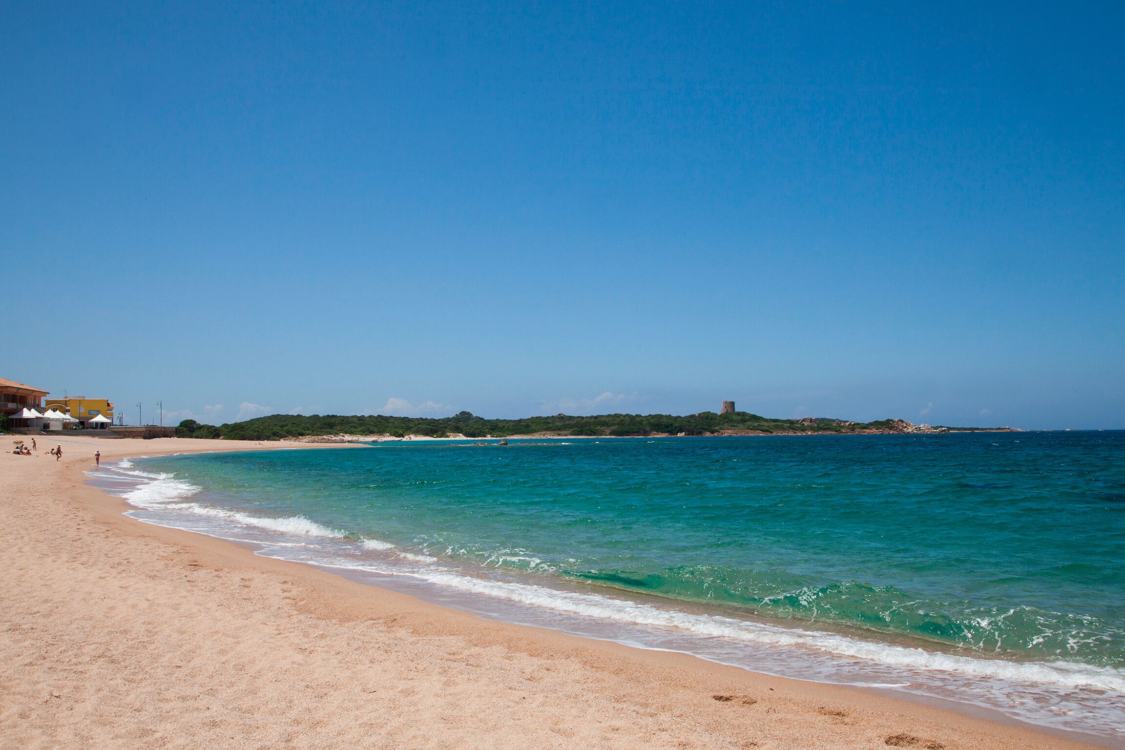 Une plage à l’eau turquoise et limpide, avec un rivage sablonneux et un bâtiment situé au loin, sur une colline.