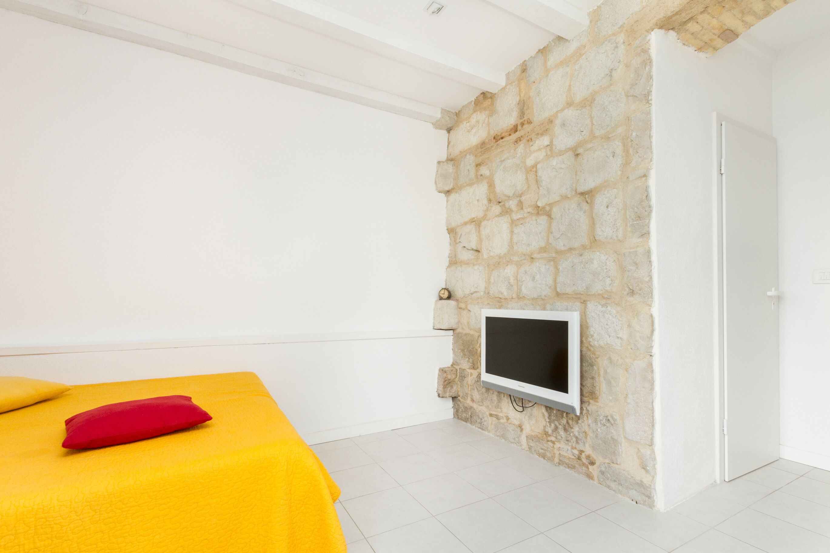 A room with a stone wall, a bed with yellow bedding and a red pillow, and a flat-screen TV mounted on the wall.