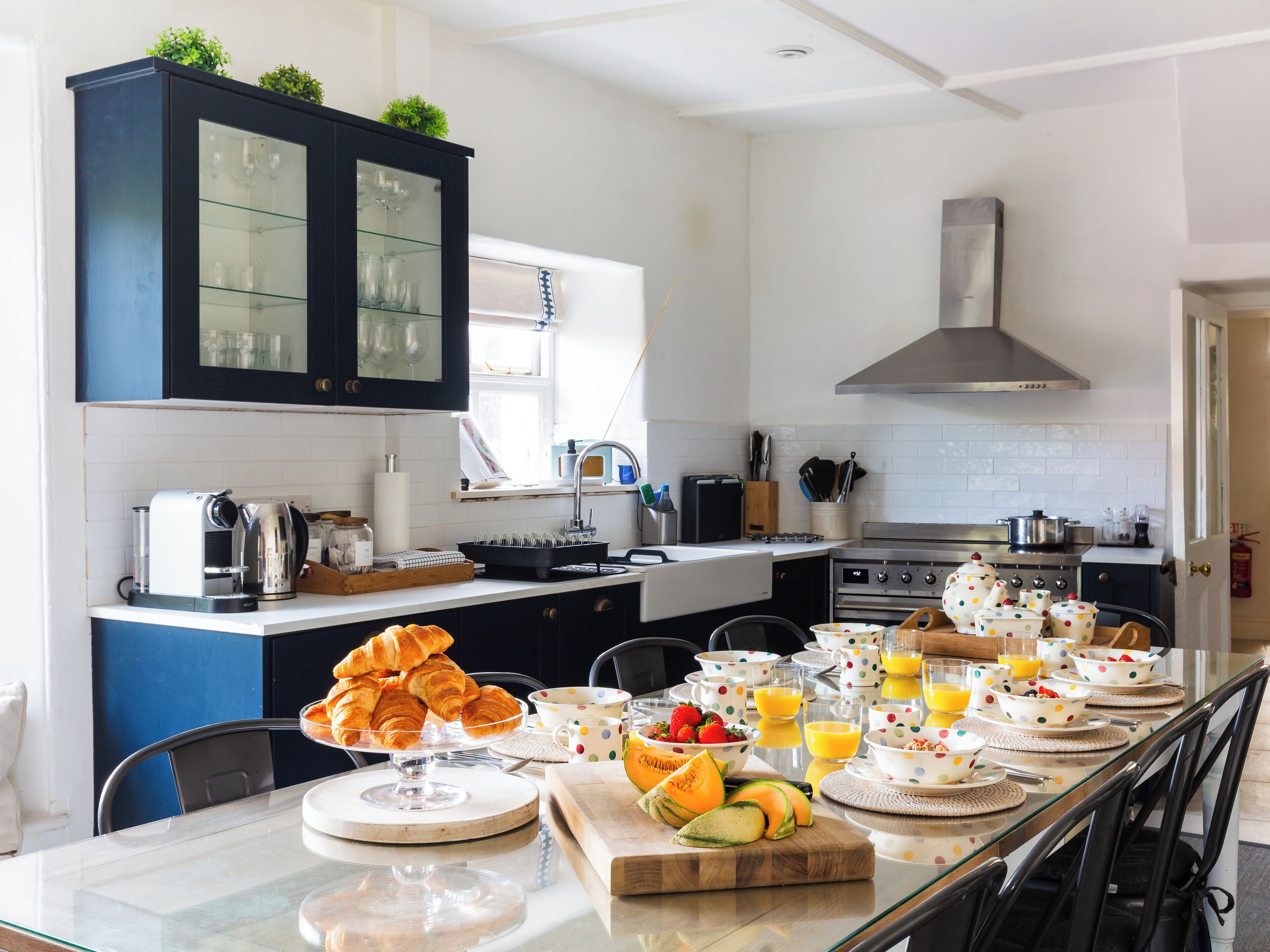 A modern kitchen with a dining area, featuring a glass-top table, a coffee machine, and a variety of breakfast items.