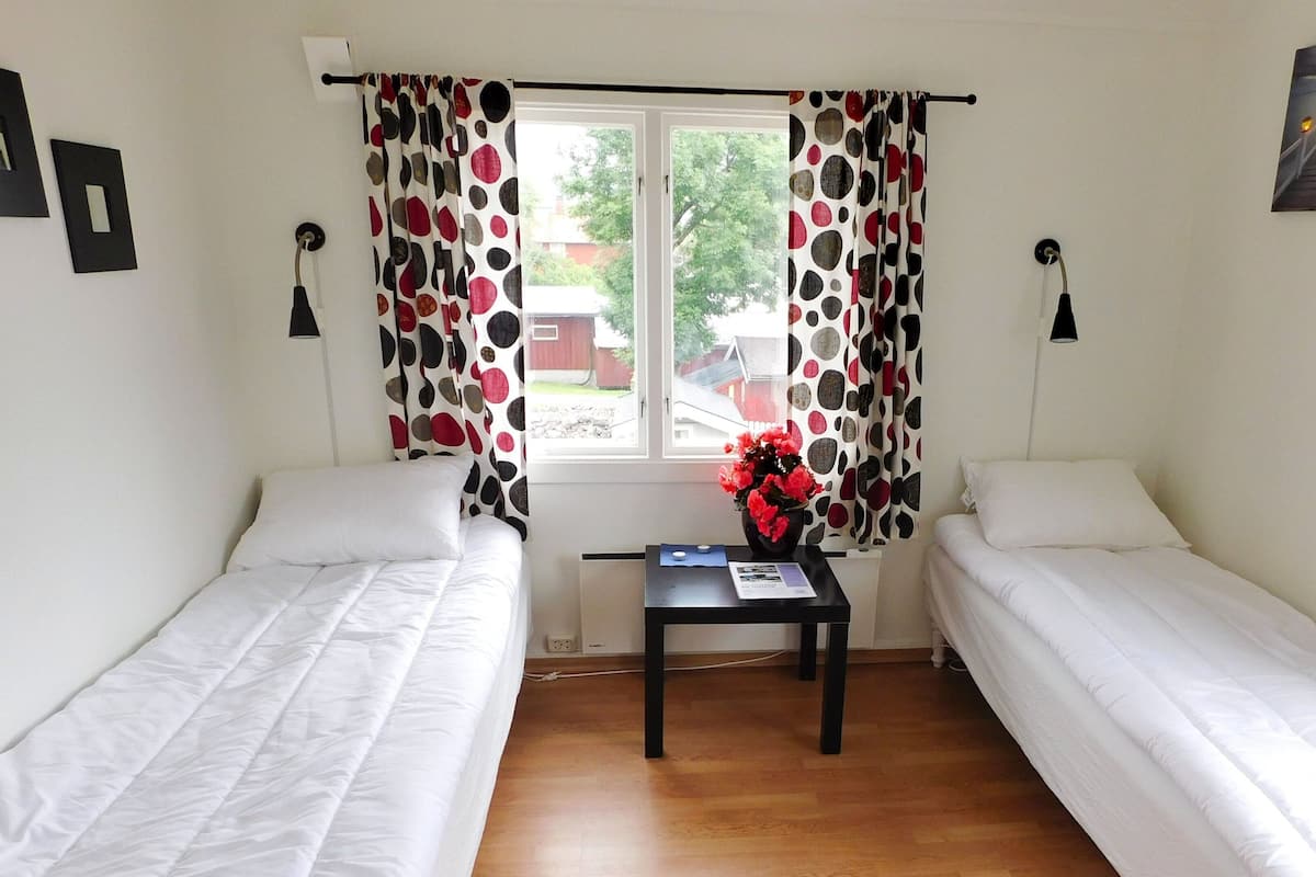 Una habitación con dos camas individuales, una mesita con un jarrón de flores y una ventana con cortinas.