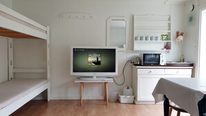 A compact room with a bunk bed, a television on a stand, a microwave, a small table with a tablecloth, and a cabinet with shelves. - Hovstø (Seljord)
