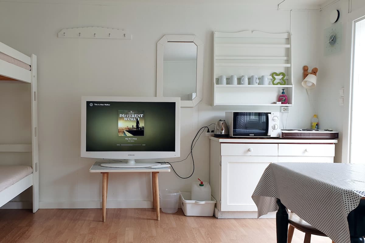 Una habitación compacta con una litera, un televisor sobre un mueble bajo, un microondas, una mesita con mantel y un armario con estantes.