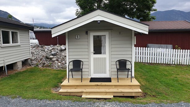 A small, single-story cabin with a white exterior and a wooden porch. There are two black chairs on the porch. - Hovstø (Seljord)
