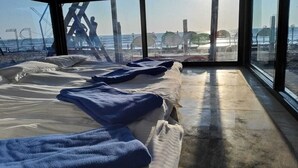 Habitación con un ventanal grande que da a la playa, con una cama, una toalla azul y vistas de sombrillas y gente.