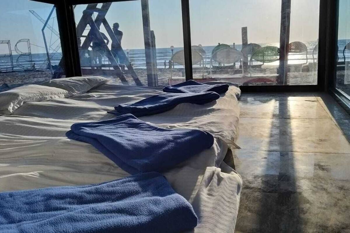 Habitación con un ventanal grande que da a la playa, con una cama, una toalla azul y vistas de sombrillas y gente.