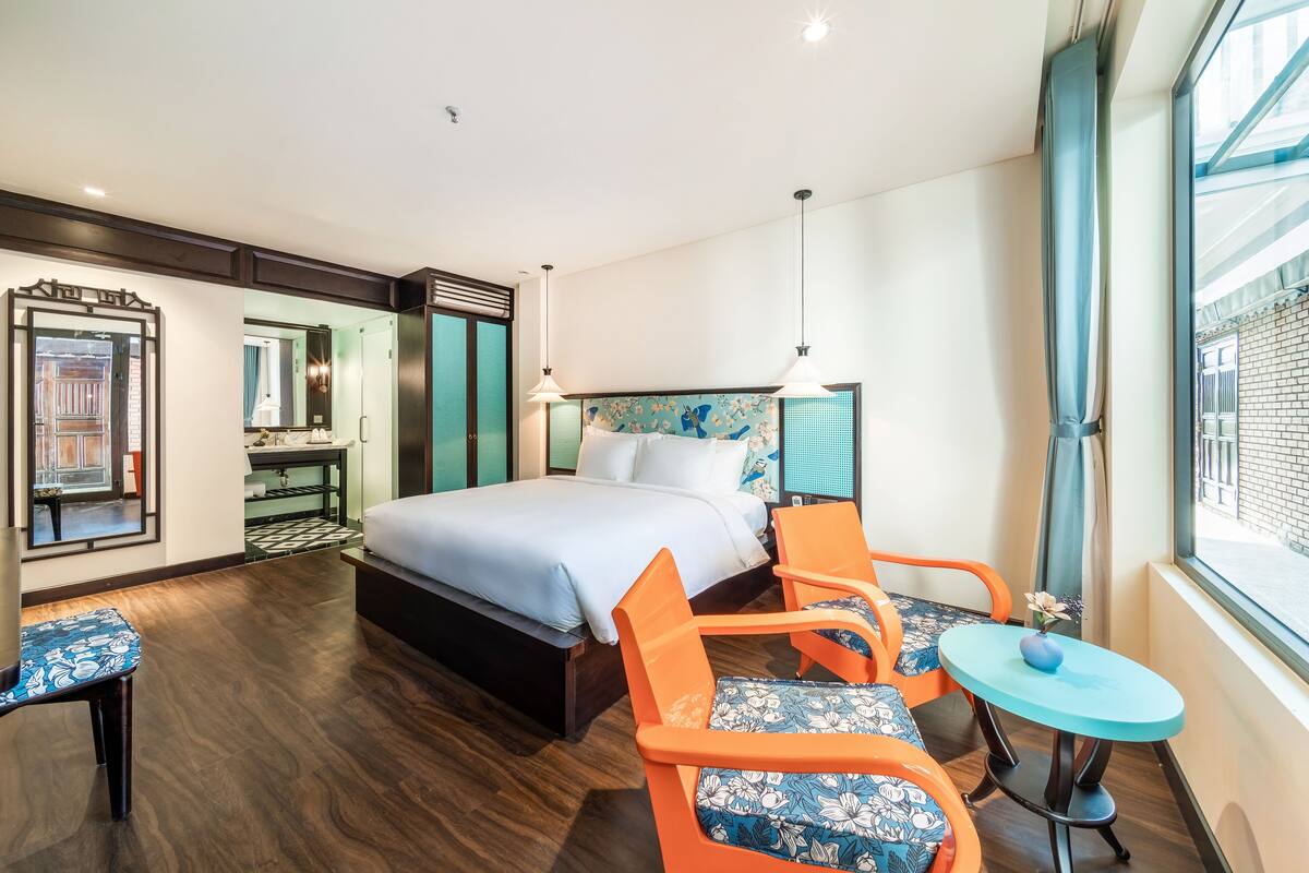 A modern hotel room with a large bed, two orange chairs, a small table, and a view of the outside through a large window.
