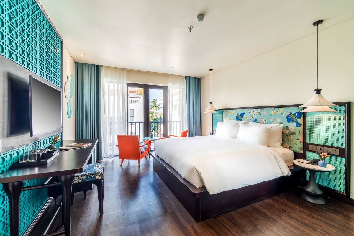A hotel room with a bed, desk, chair, TV, and a view of the outside.