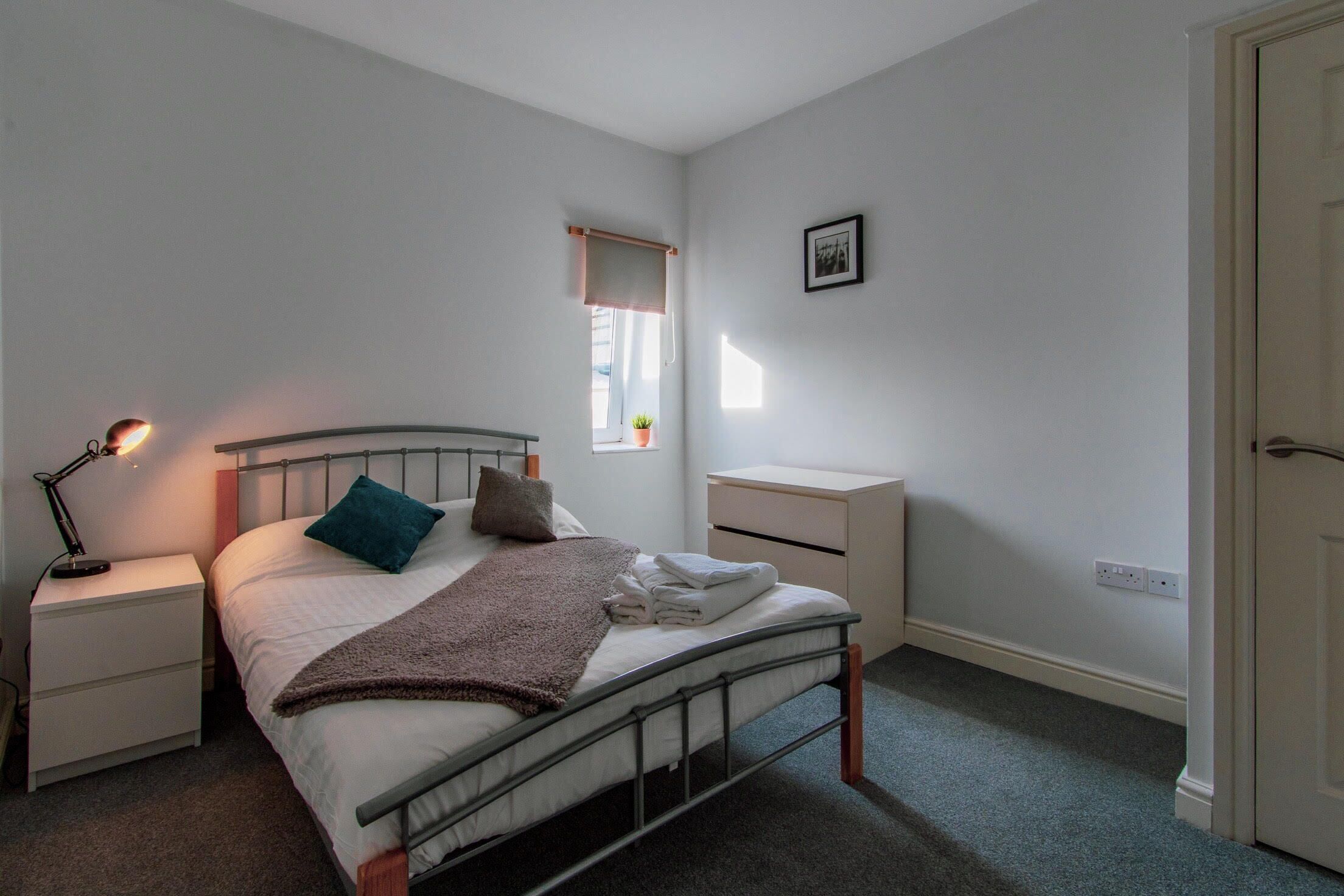 A bedroom with a single bed, a bedside table with a lamp, a small window with a plant, and a door.