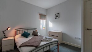 A bedroom with a single bed, a bedside table with a lamp, a small window with a plant, and a door. - 70 Elm Street (Cardiff)