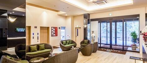 A modern hotel lobby with wicker furniture, a wooden floor, and large windows offering a view of the outdoors.