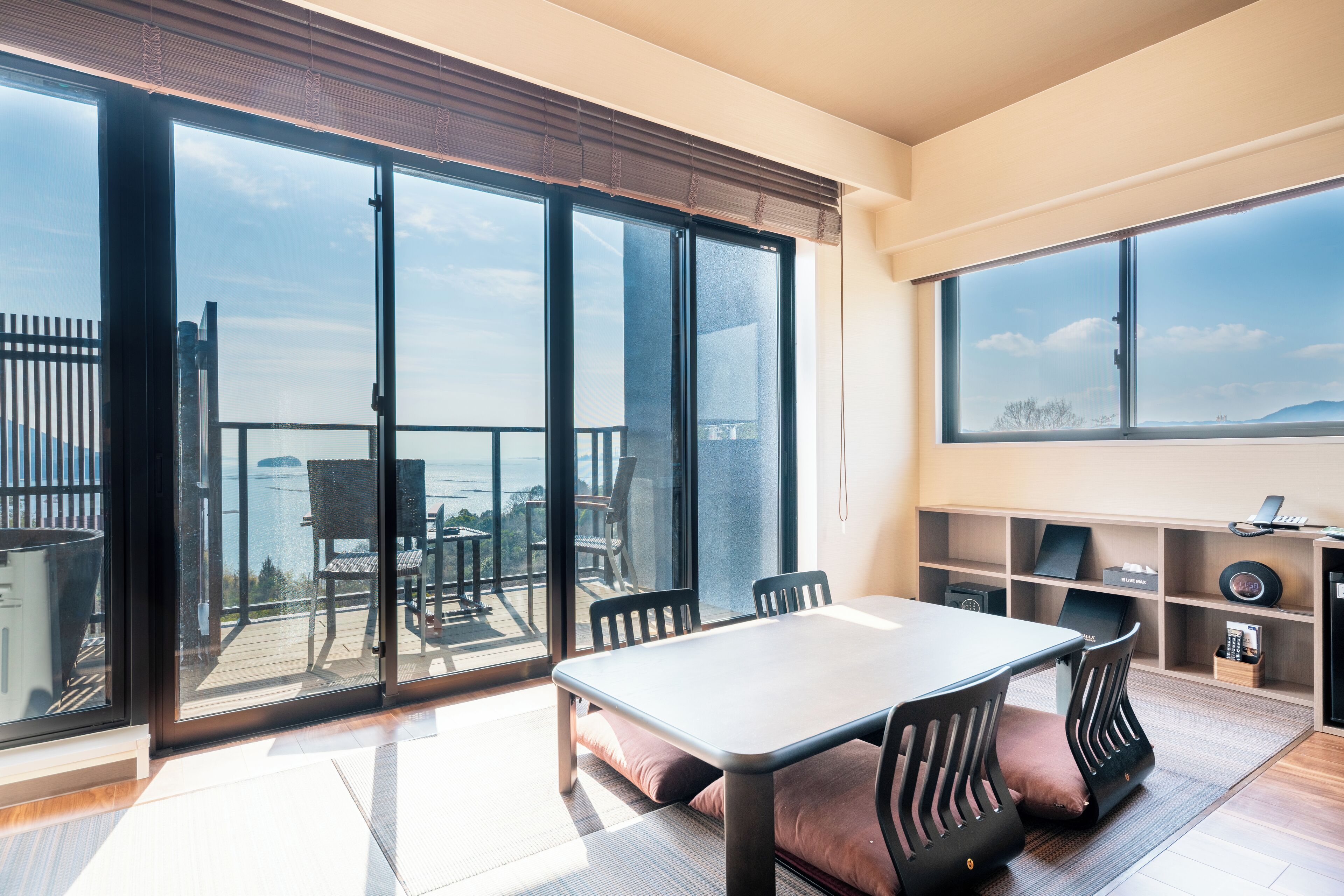 A modern living room with a large sliding glass door leading to a balcony, a dining area with a table and chairs, and a shelving unit with various items.