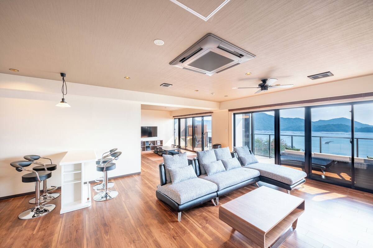 A modern living room with a sectional sofa, a coffee table, and a bar area with stools. There is a large window offering a view of the sea and mountains.