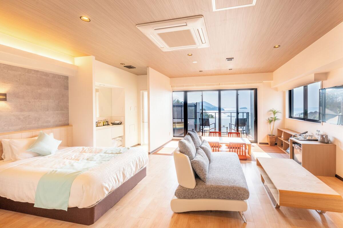 A modern hotel room with a large bed, a sofa, a dining table, and a balcony with a view of the sea.
