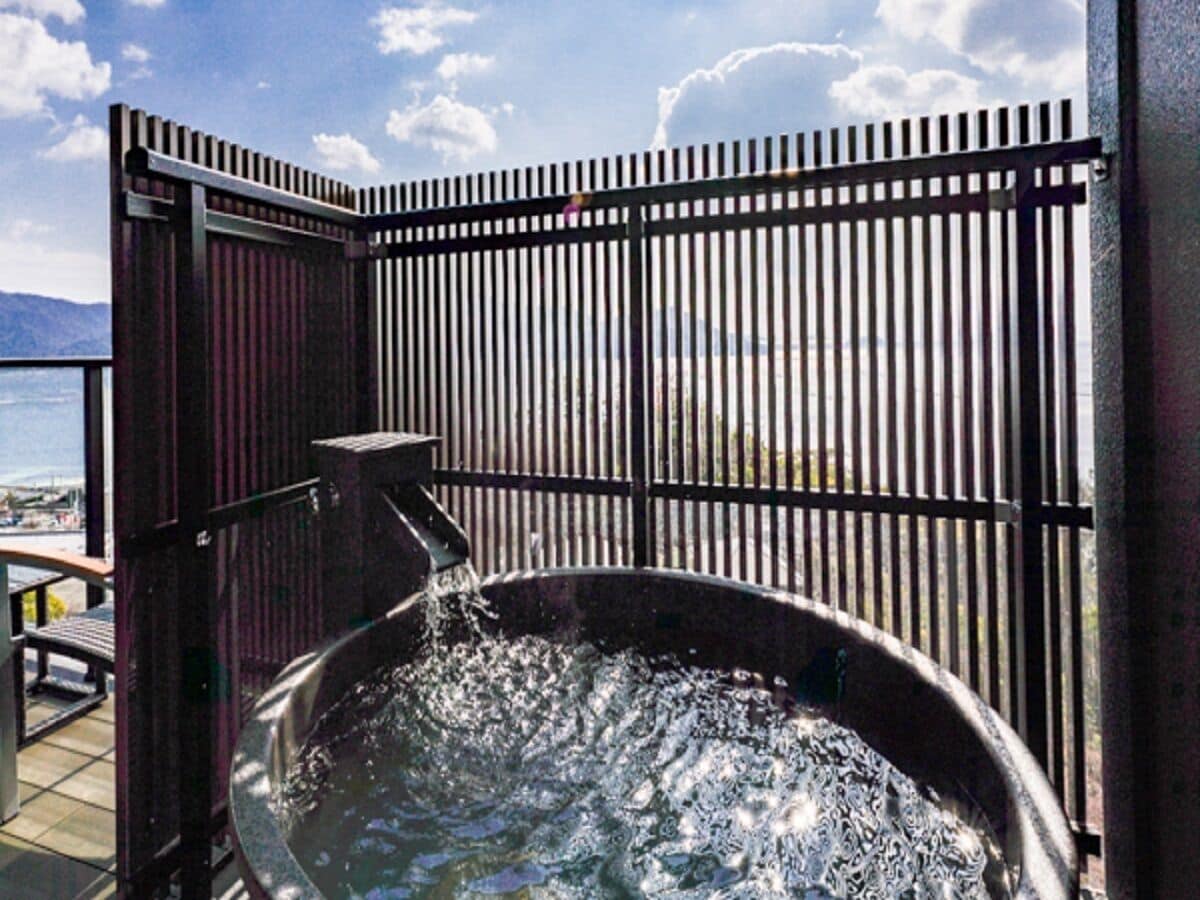 A hot tub with water flowing from a faucet, enclosed by a metal fence with a view of the sea in the background.