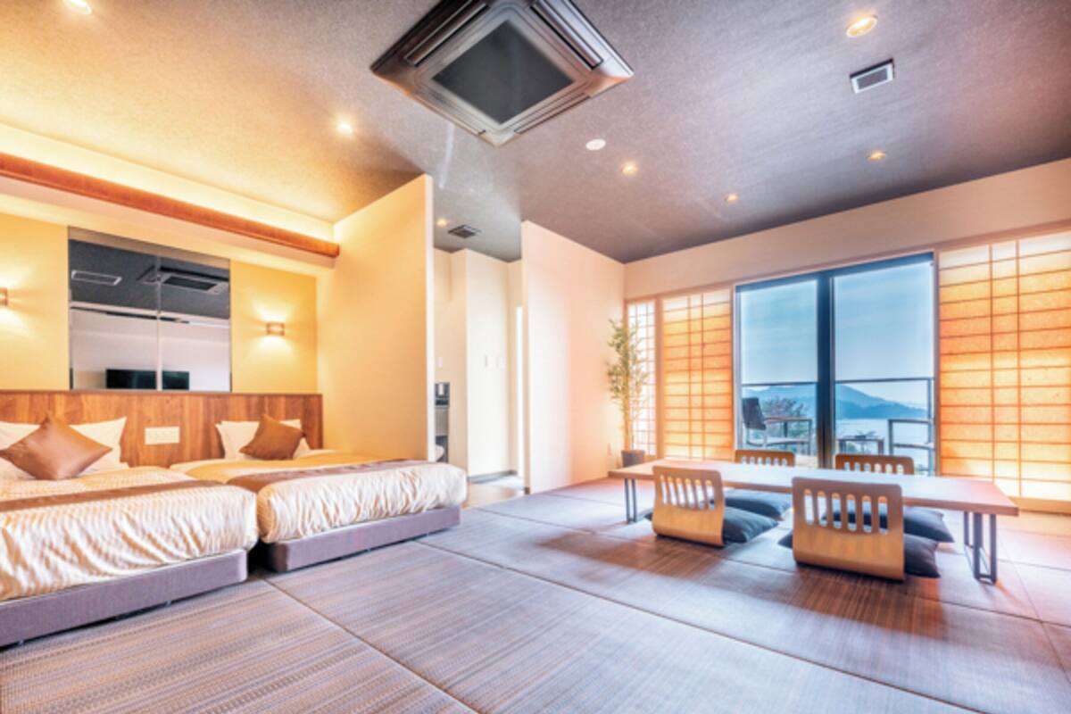 A modern hotel room with a large bed, a seating area, and a view of the outdoors through large windows.