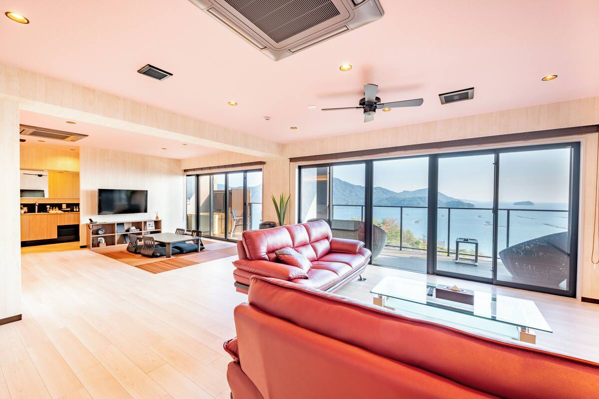 A modern living room with a leather sofa, a flat-screen TV, and a large window offering a view of the sea.