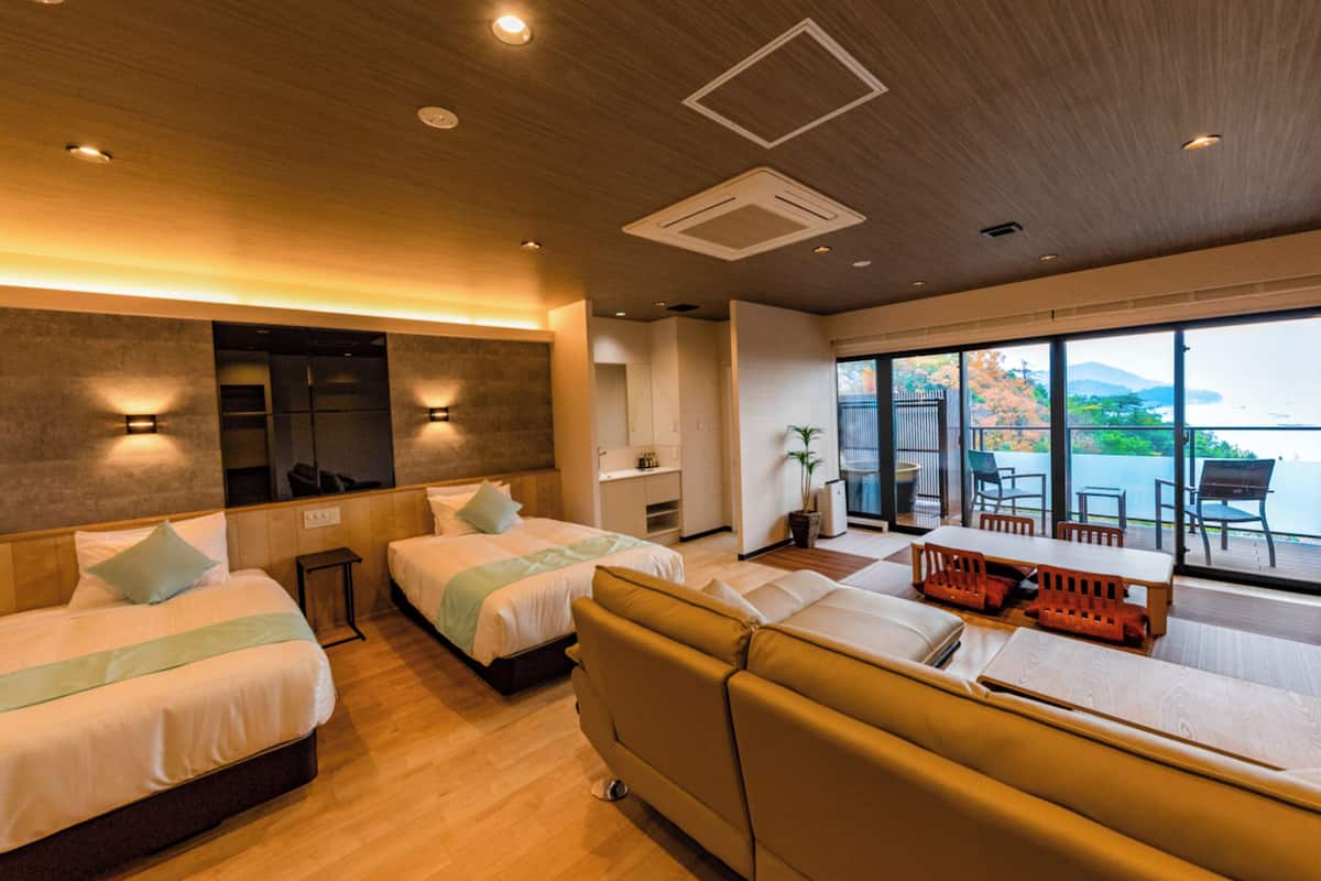 A modern hotel room with a large bed, a sofa, a dining table, and a view of the sea.