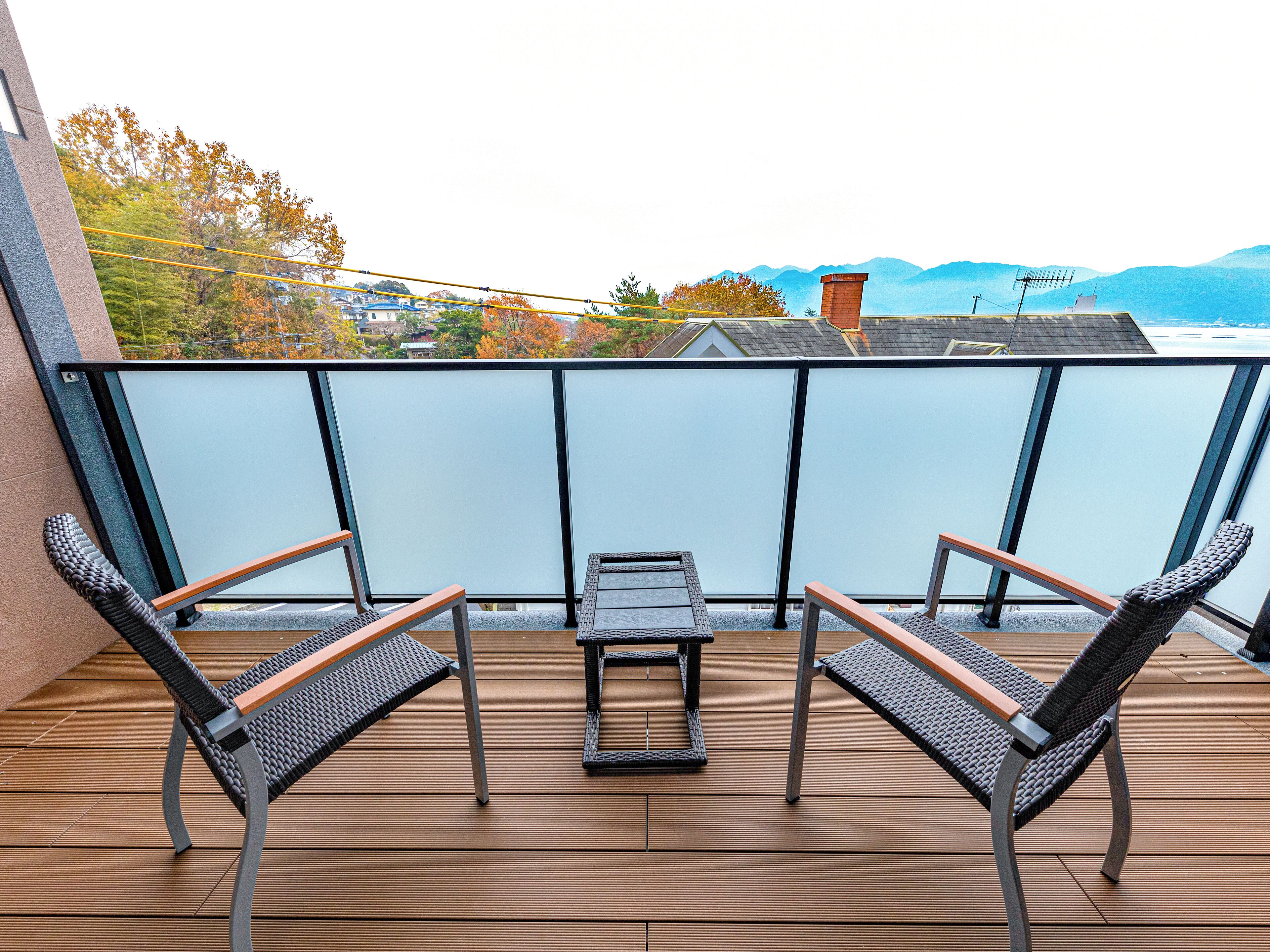 A balcony with two wicker chairs and a small table, overlooking a scenic view with mountains and autumn trees.
