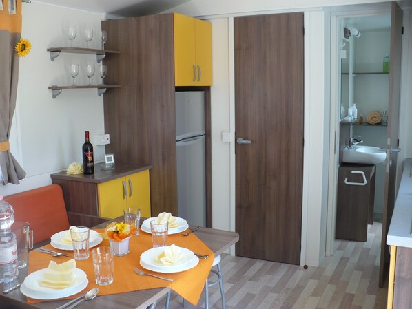 A compact kitchen with a refrigerator, a sink, and a dining area set with plates, glasses, and a bottle of wine. - Victoria Mobilehome Camping Union Lido Vacanze (Cavallino-Treporti)
