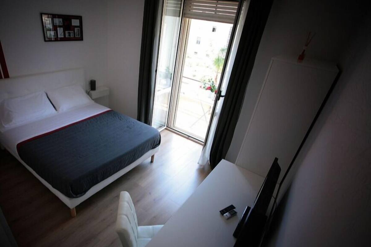 A bedroom with a bed, a desk, and a chair. There is a window with curtains and a view of the outside.