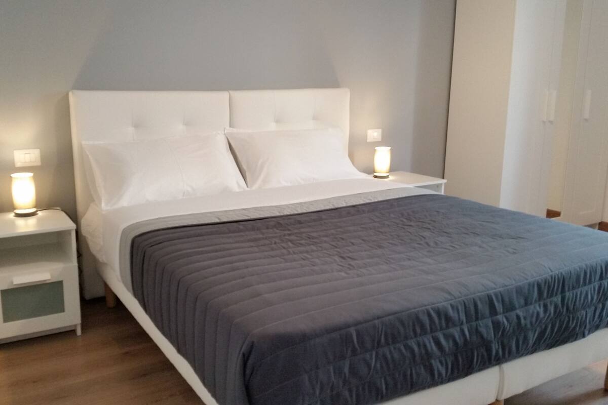 A neatly made bed with a grey duvet, white pillows, and a white headboard. There are two bedside tables with lamps on either side of the bed.
