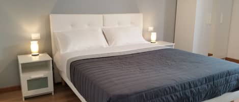 A neatly made bed with a grey duvet, white pillows, and a white headboard. There are two bedside tables with lamps on either side of the bed.