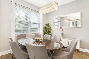 Un coin repas avec une table ronde en verre, des chaises à revêtement gris, une plante en pot et un grand miroir accroché au mur.