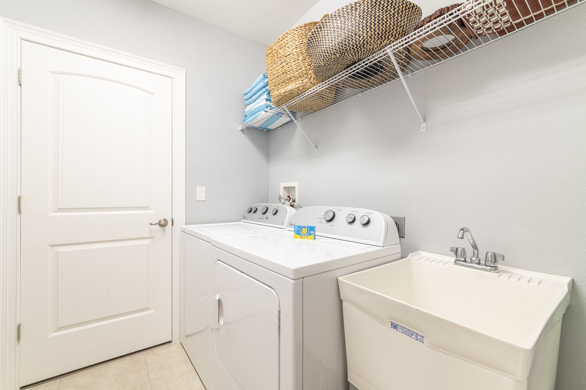 A laundry room with a white door, a washing machine, a dryer, a sink, and a basket rack with folded clothes.