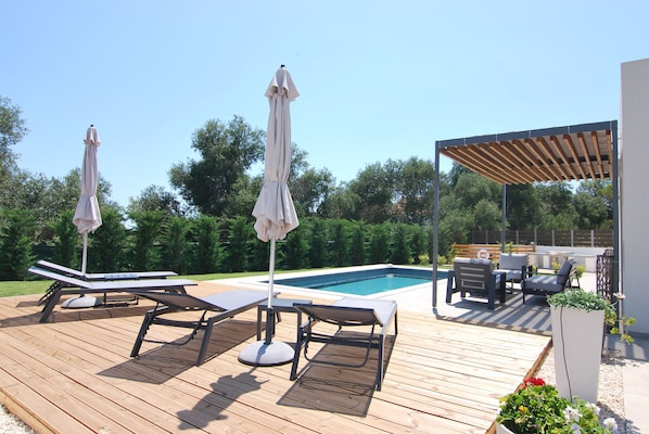 Una terraza de madera con sillones de descanso y sombrillas, una piscina y un área de estar cubierta.