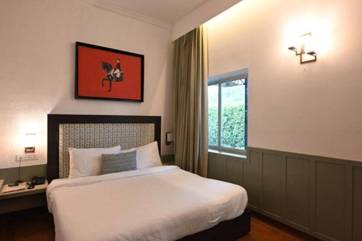Una habitación de hotel con cama, mesita de noche, ventana con cortinas y lámpara de pared.