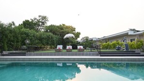A swimming pool with lounge chairs and umbrellas, surrounded by greenery and a building. - Status Club Resort (Kanpur)