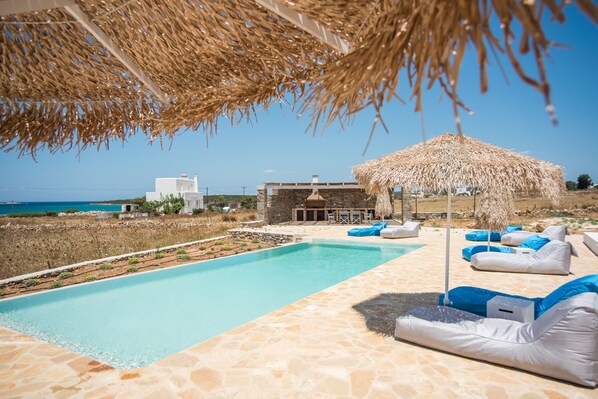 A pool area with lounge chairs and a thatched umbrella, a stone pathway, and a view of the sea and buildings in the background.