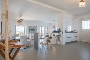 Un vaste espace de vie comprenant une table à manger en bois, des canapés blancs et un coin cuisine avec des tabourets.
