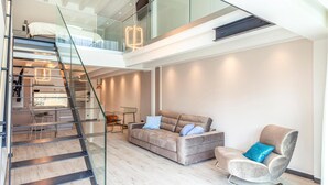 A modern interior with a staircase leading to a loft bedroom, a living area with a sofa and armchair, and a dining area with a glass table and chairs. - Prora Solitaire Avida Loft12 (Binz)