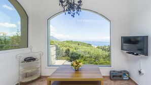 Een kamer met een groot boogvenster met uitzicht op groen en de zee. Er staat een houten tafel met een bloevas, een aan de muur bevestigde televisie en een metalen plank met een tas.
