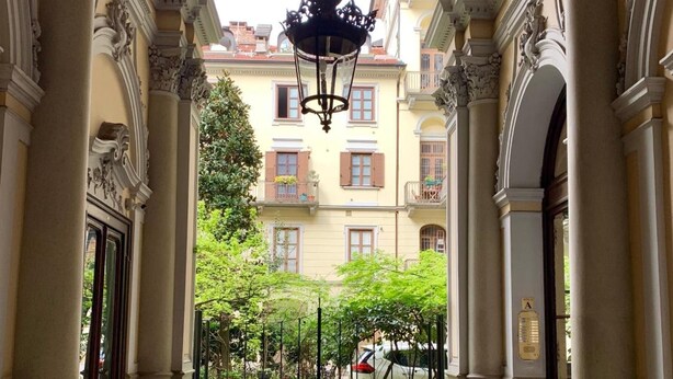 Un couloir orné de colonnes décoratives, un lustre et une entrée fermée par un portail menant à un bâtiment doté de balcons.