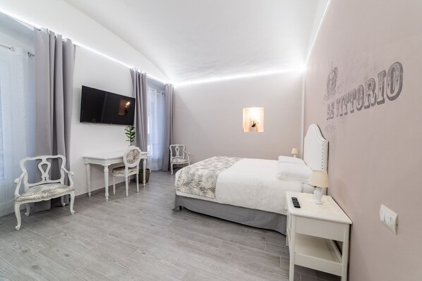 A hotel room with a bed, a desk, a chair, a TV, and a wall-mounted light.