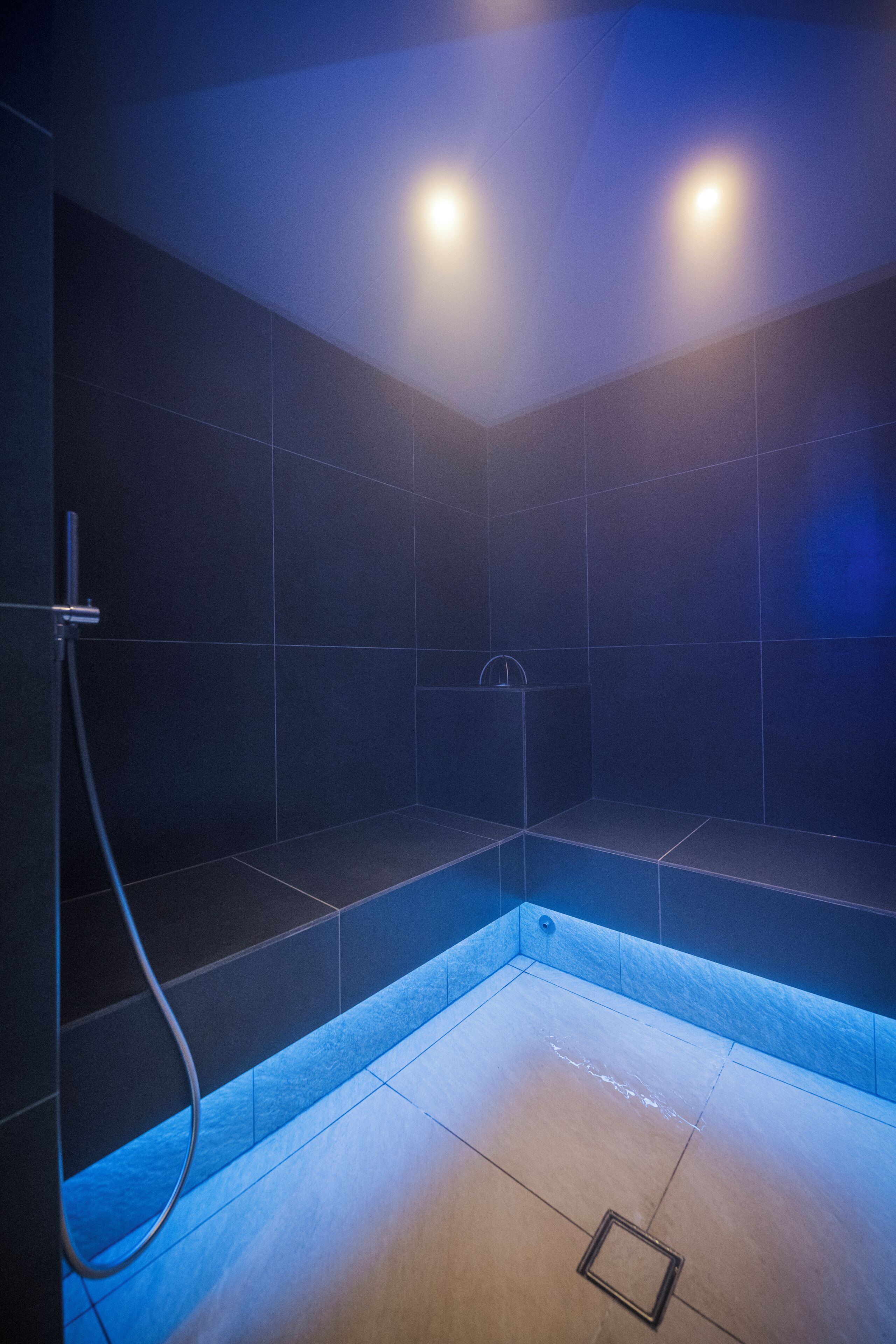 A modern shower room with dark tiles, a visible drain, and a handheld showerhead.